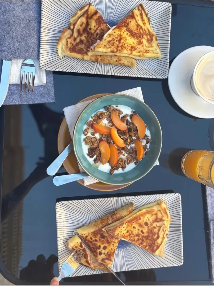 Un tavolo apparecchiato con due piatti di crepes, una scodella di frutta e cereali e un succo