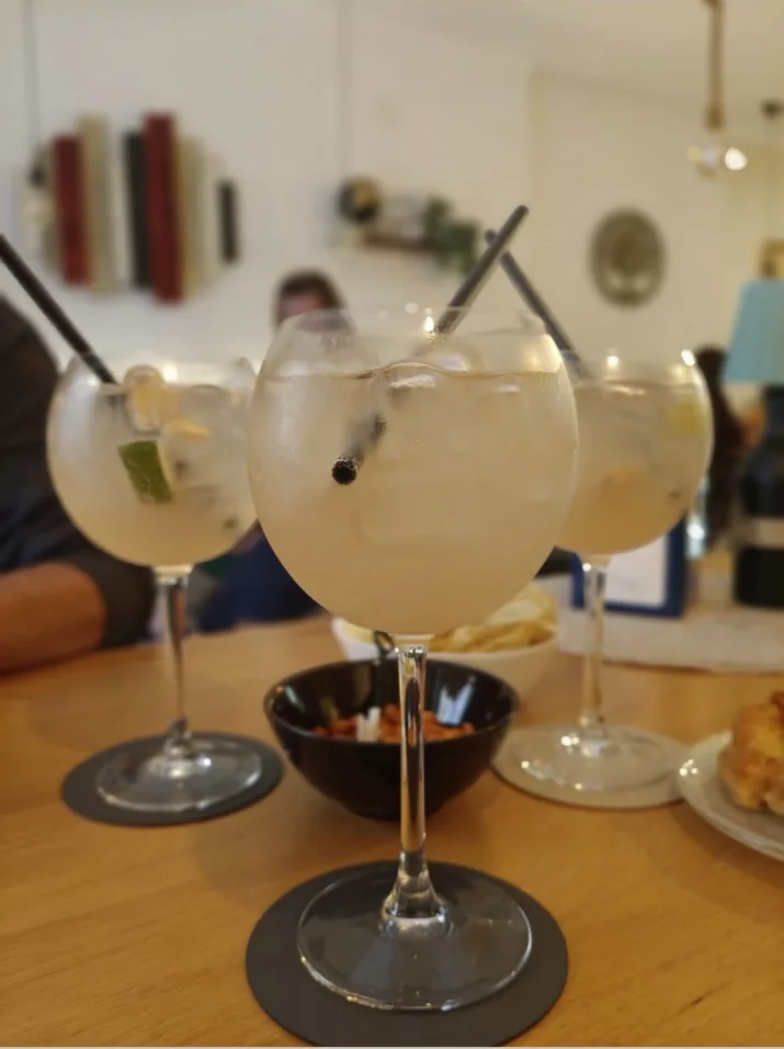 Tre calici di gin tonic su un tavolo di legno chiaro, sullo sfondo la sala luminosa con le mensole e le lampade