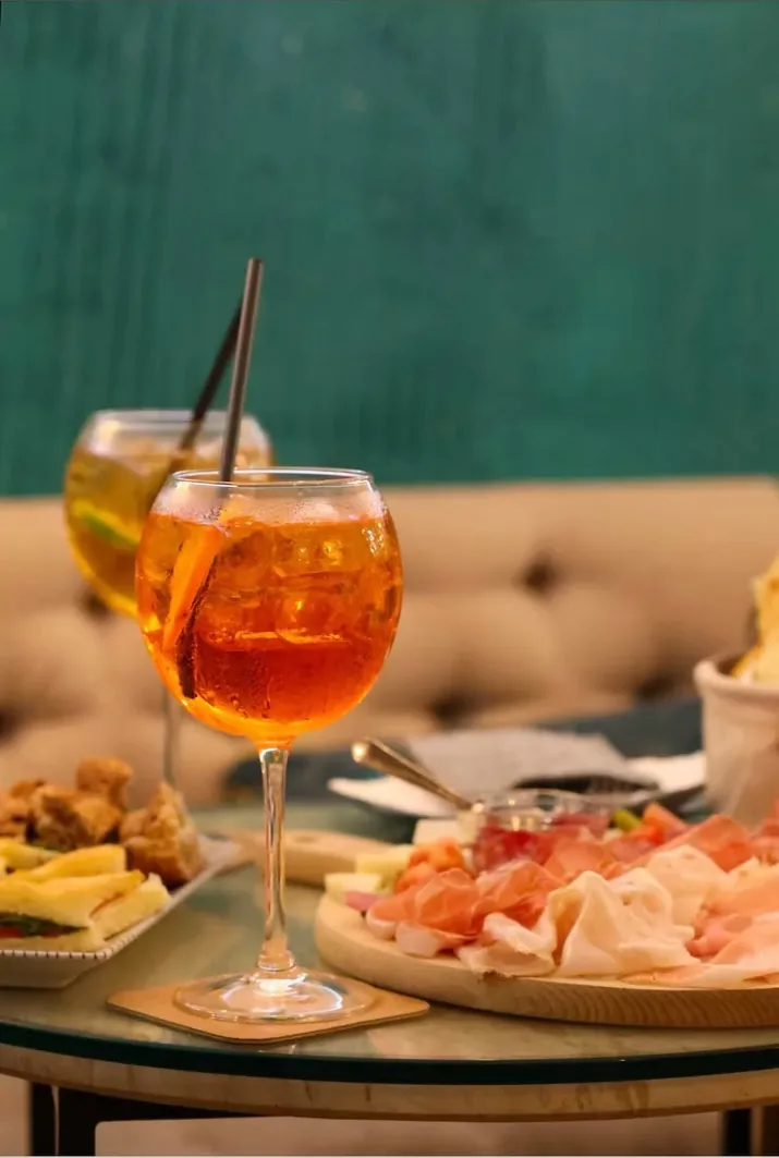 Un calice di spritz con una fetta d'arancia e un tagliere di salumi e formaggi su un tavolino di vetro, davanti alla parete verde bottiglia con le linee dorate della sala